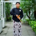 A man in traditional attire stands confidently in a lush garden setting.