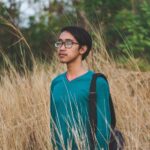 Portrait of a young man in a blue sweater and glasses, standing in tall grass outdoors.