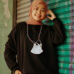 Portrait of an Asian woman in black sweater and hijab, standing against a painted wall in Cirebon.