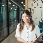 Asian woman smiling with a clipboard in hand, standing in a modern office environment.