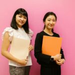 Two professional women smiling and holding folders against a pink background. Ideal for business and corporate themes.